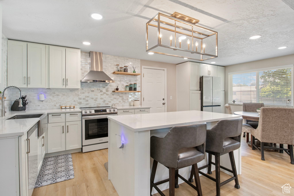 Kitchen featuring wall chimney range hood, stainless steel range with electric stovetop, freestanding refrigerator, a kitchen island, and light countertops