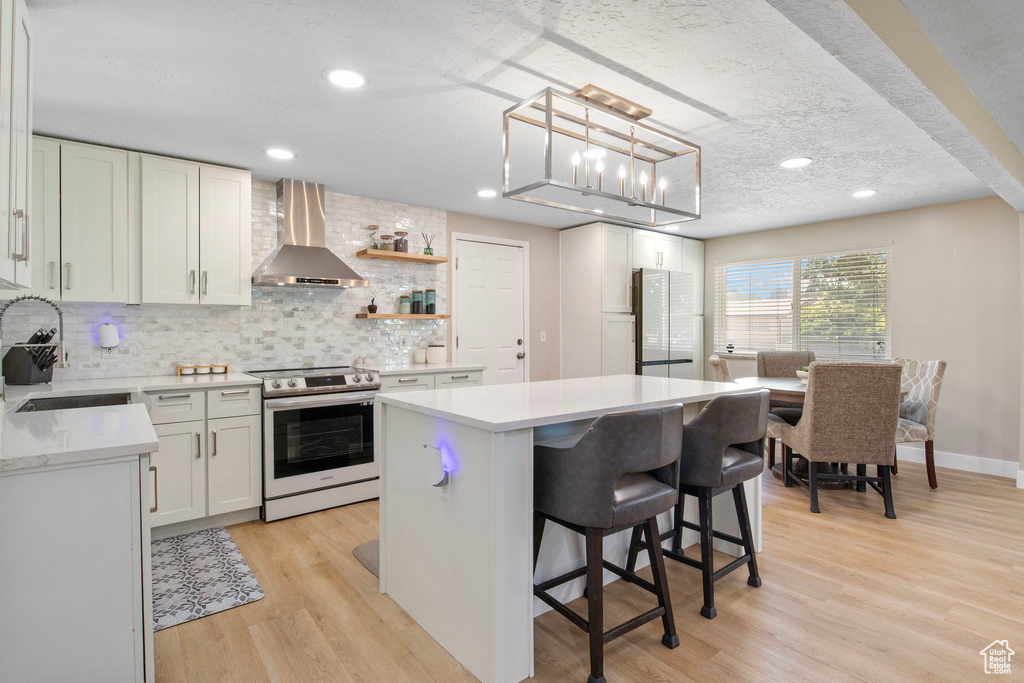 Kitchen with stainless steel electric stove, wall chimney range hood, freestanding refrigerator, a center island, and light countertops