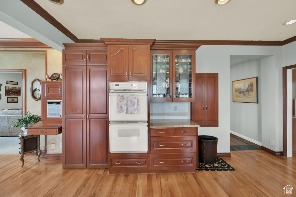 Kitchen with double oven, light wood finished floors, decorative backsplash, glass insert cabinets, and crown molding