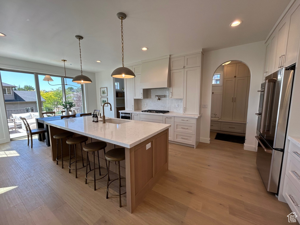 Kitchen with custom exhaust hood, stainless steel appliances, light wood-style floors, recessed lighting, and a center island with sink