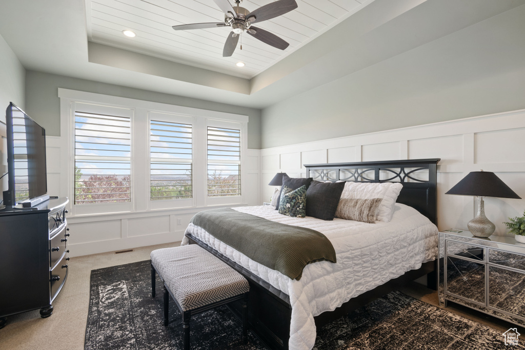 Bedroom with a decorative wall, a wainscoted wall, a raised ceiling, light colored carpet, and a ceiling fan