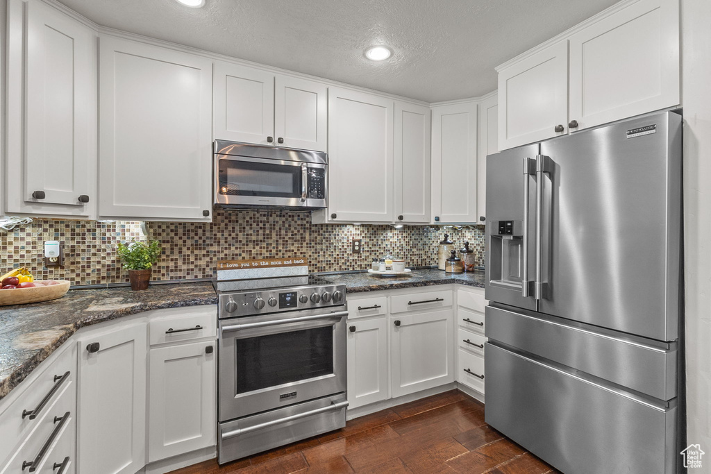 Kitchen featuring appliances with stainless steel finishes, dark wood-style flooring, white cabinets, tasteful backsplash, and dark stone countertops
