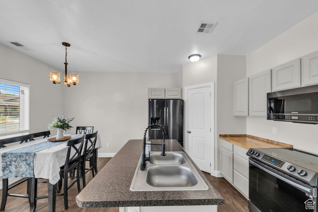 Kitchen with appliances with stainless steel finishes, dark wood-style floors, a center island with sink, a chandelier, and dark countertops