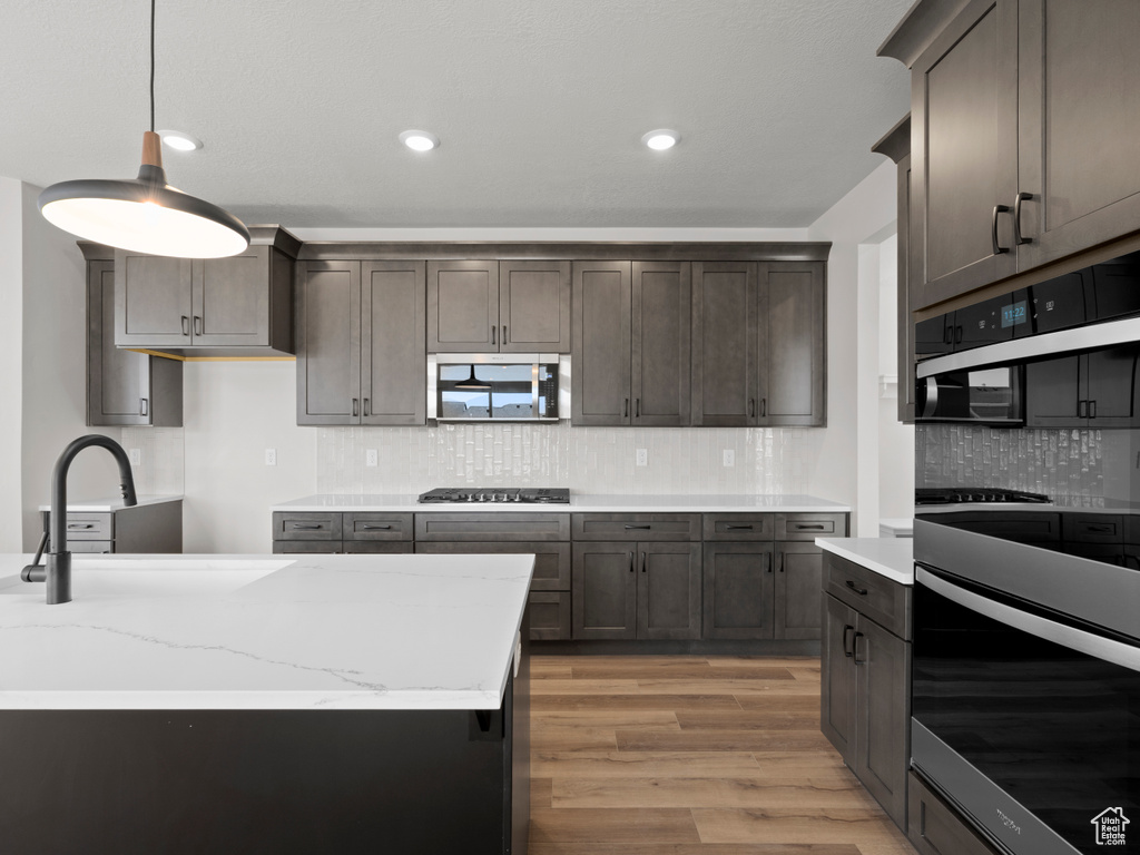 Kitchen featuring appliances with stainless steel finishes, light wood finished floors, decorative backsplash, decorative light fixtures, and light stone counters