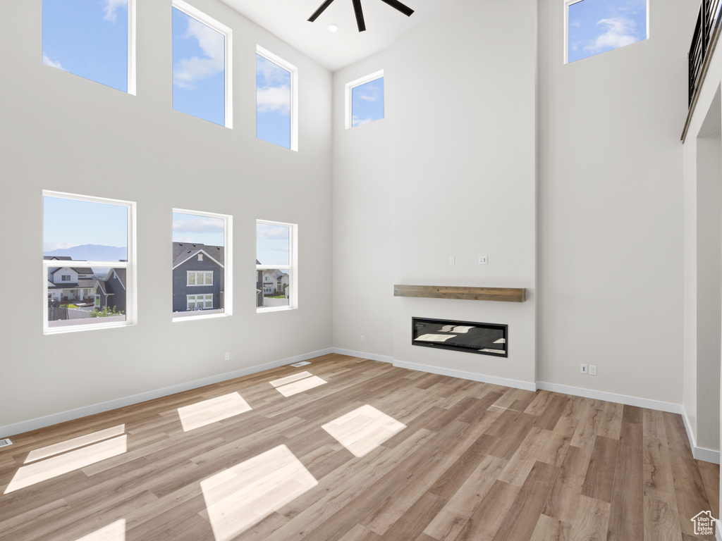 Unfurnished living room with a towering ceiling, a ceiling fan, light wood-style flooring, and a glass covered fireplace