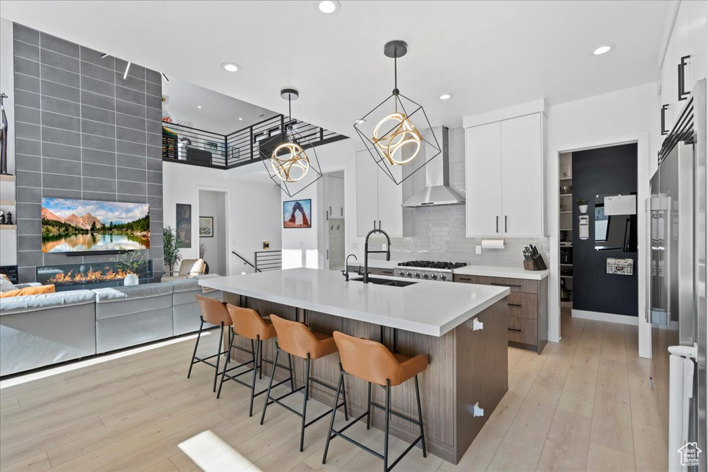 Kitchen with wall chimney range hood, a breakfast bar area, an island with sink, light countertops, and white cabinets