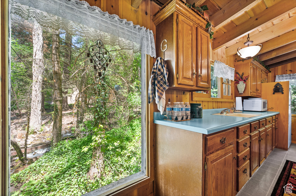 Kitchen with white microwave, wooden walls, brown cabinets, a wood ceiling with exposed beams, and light countertops