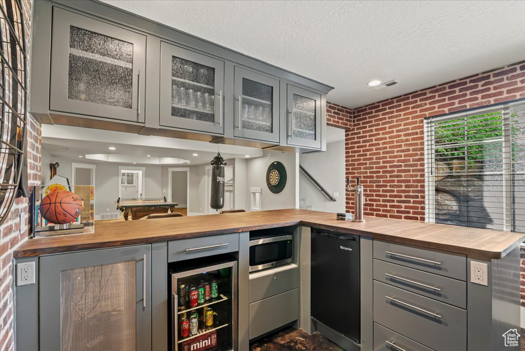 Indoor wet bar featuring brick wall, stainless steel microwave, beverage cooler, a textured ceiling, and dishwashing machine