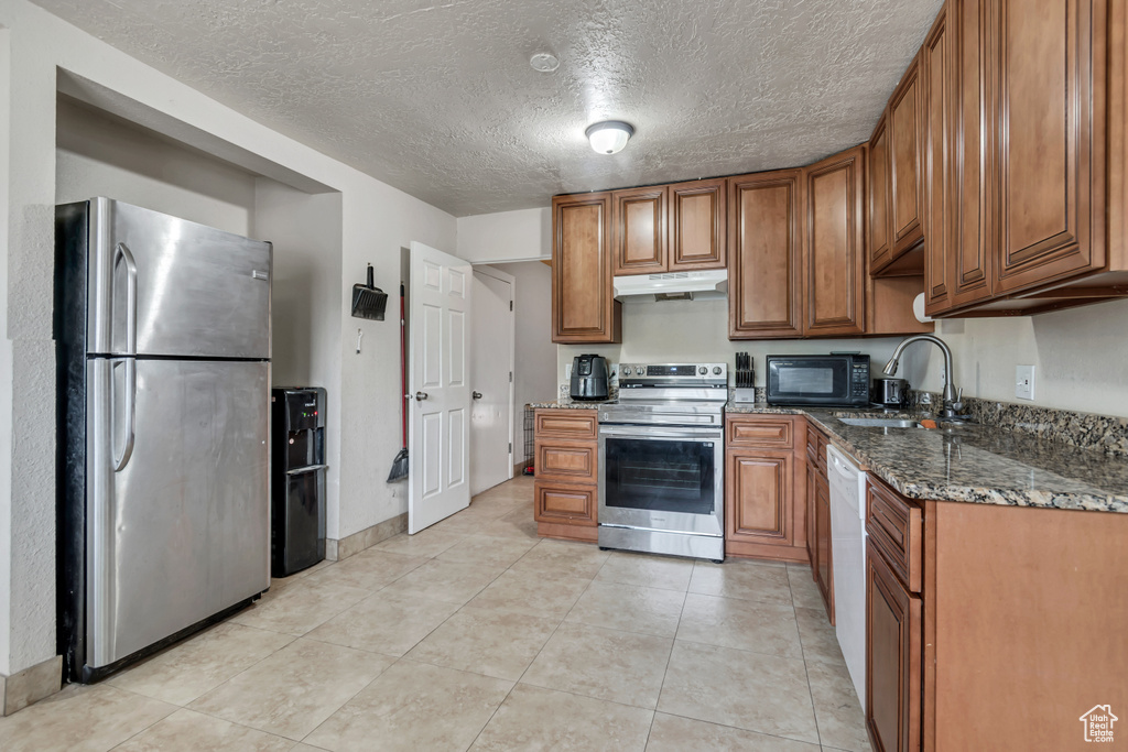 Kitchen featuring appliances with stainless steel finishes, under cabinet range hood, brown cabinets, dark stone counters, and a textured ceiling
