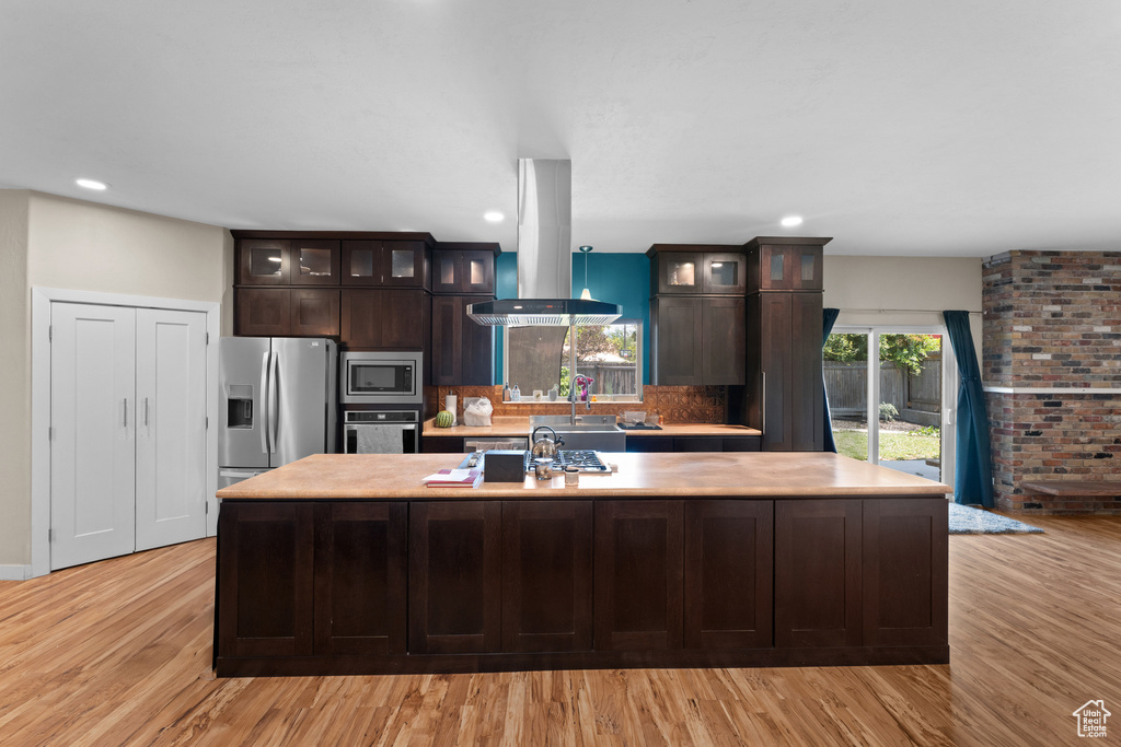 Kitchen with appliances with stainless steel finishes, island exhaust hood, light countertops, a center island with sink, and dark brown cabinets