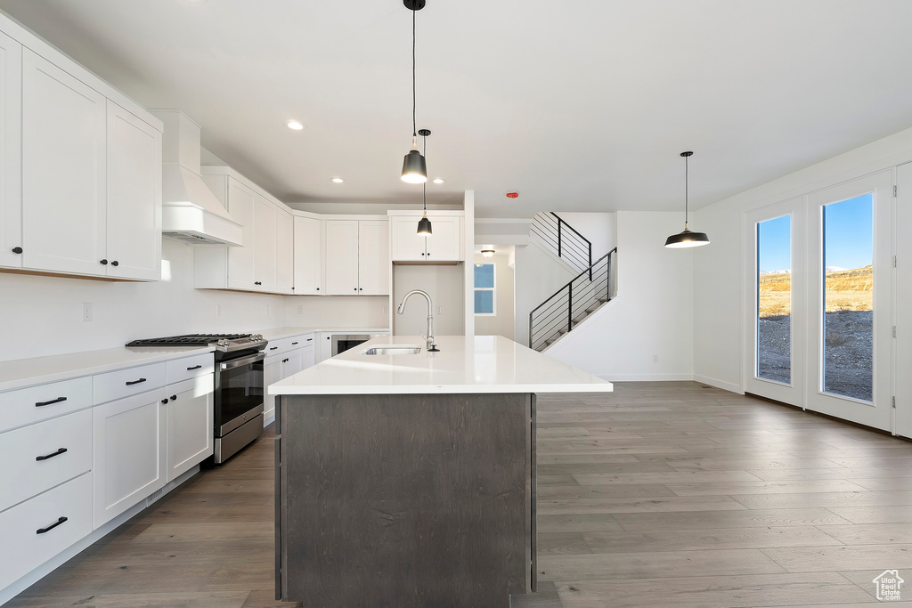 Kitchen with stainless steel range with gas cooktop, premium range hood, wood finished floors, a kitchen island with sink, and white cabinets