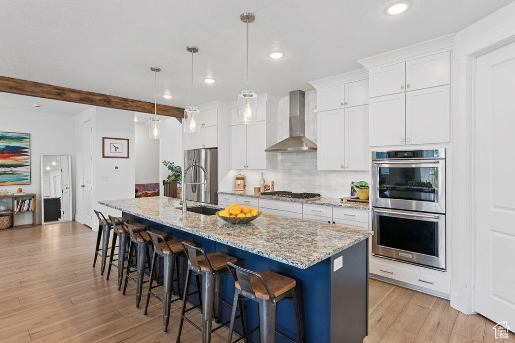 Kitchen featuring wall chimney exhaust hood, stainless steel appliances, white cabinets, light wood-style floors, and a center island with sink