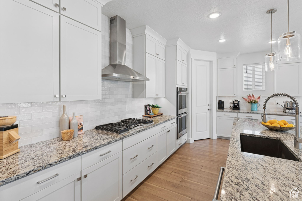 Kitchen featuring wall chimney exhaust hood, stainless steel appliances, white cabinetry, backsplash, and pendant lighting