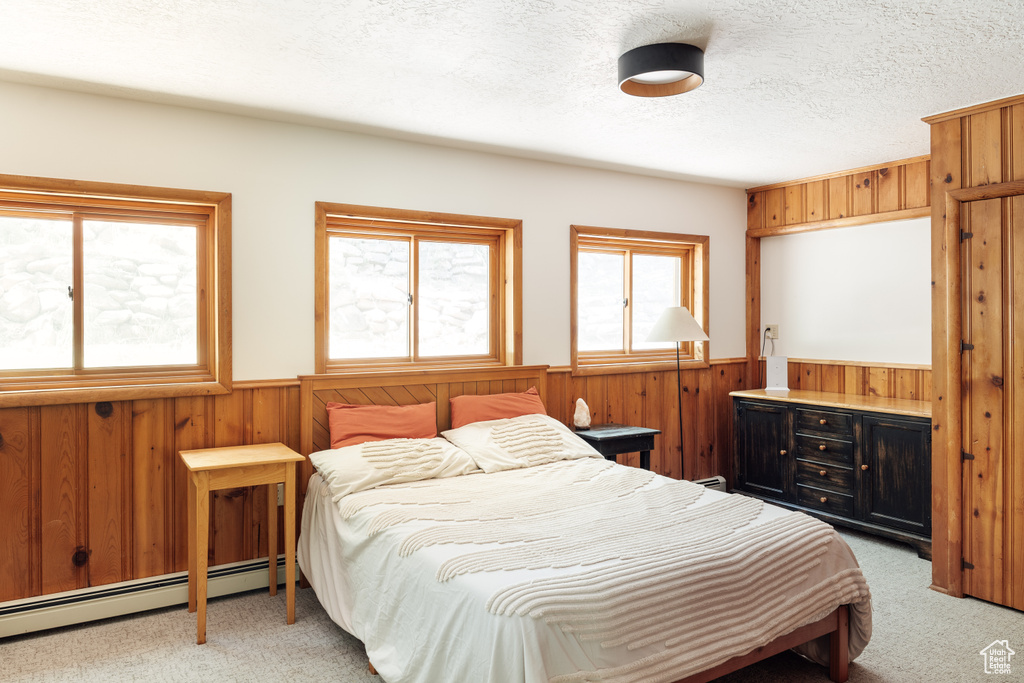 Bedroom with a baseboard radiator, wainscoting, light colored carpet, wood walls, and a textured ceiling