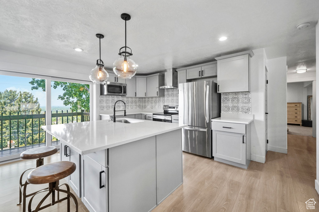 Kitchen with appliances with stainless steel finishes, light countertops, tasteful backsplash, light wood-style floors, and wall chimney range hood