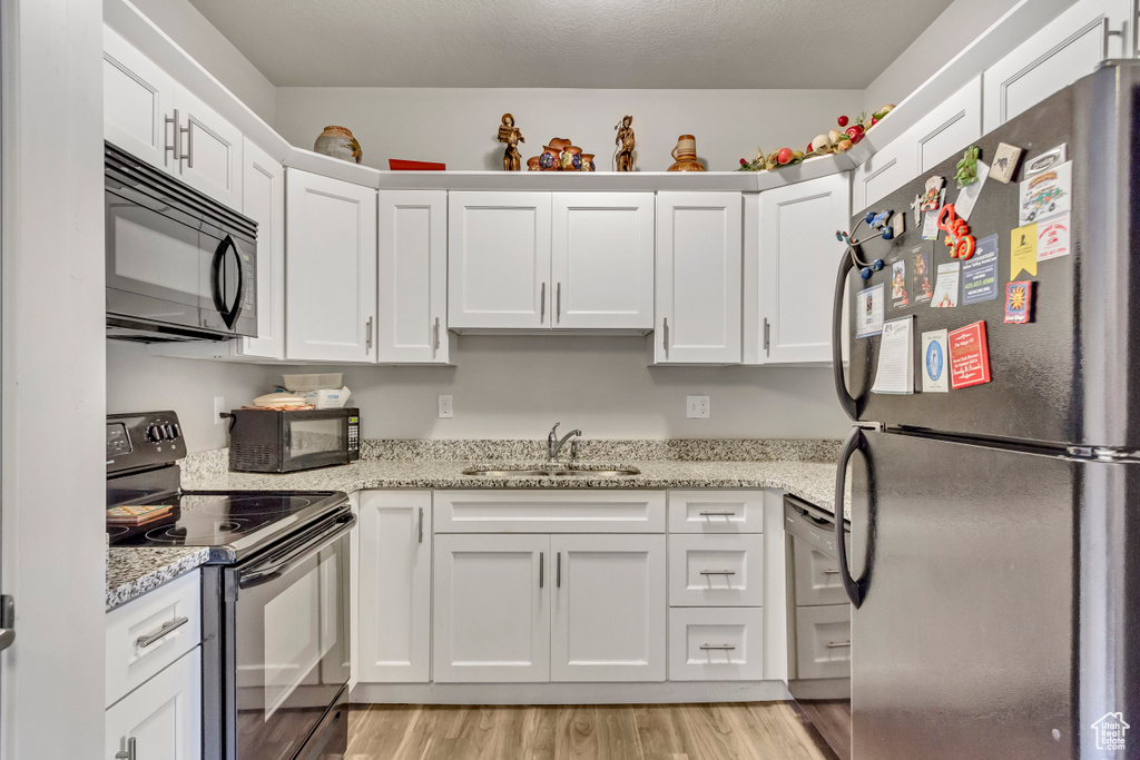 Kitchen with black appliances, a sink, white cabinets, light wood-type flooring, and light stone countertops