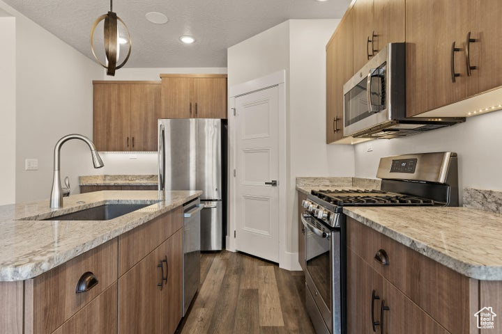 Kitchen featuring appliances with stainless steel finishes, dark wood-style floors, light stone counters, decorative light fixtures, and a kitchen island with sink
