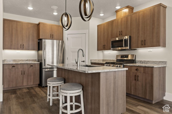 Kitchen featuring stainless steel appliances, dark wood-style floors, a kitchen bar, hanging light fixtures, and light stone countertops