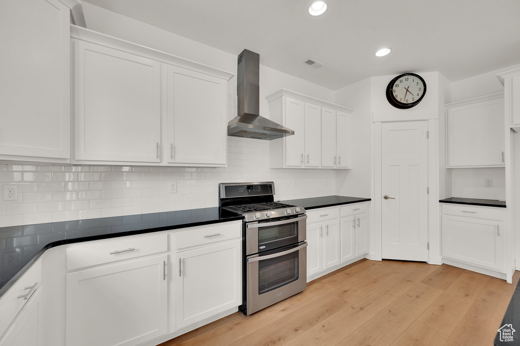 Kitchen with wall chimney exhaust hood, double oven range, white cabinets, light wood-style flooring, and dark countertops