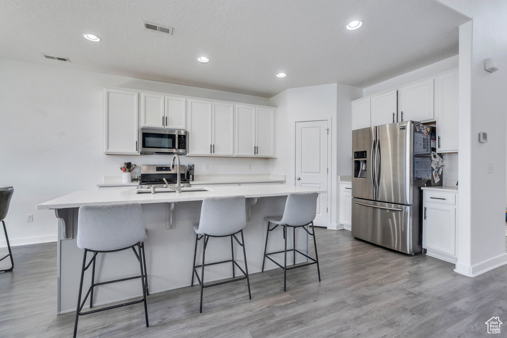 Kitchen featuring stainless steel appliances, white cabinetry, light countertops, light wood finished floors, and a kitchen island with sink