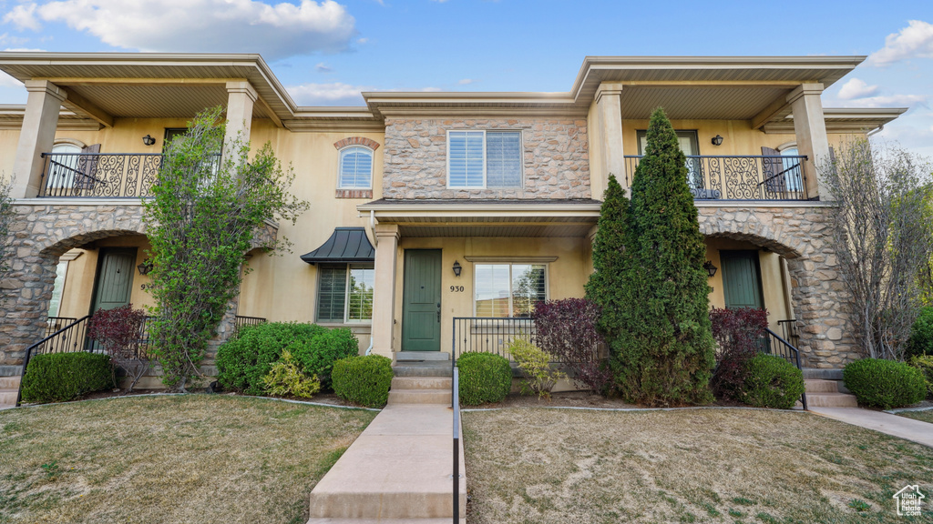 View of front of house featuring a balcony, stucco siding, stone siding, and a front yard