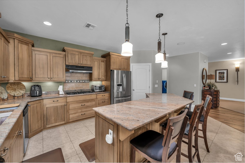 Kitchen with appliances with stainless steel finishes, under cabinet range hood, light tile patterned flooring, a kitchen island, and light stone countertops