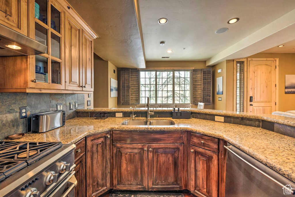 Kitchen with appliances with stainless steel finishes, under cabinet range hood, light stone countertops, recessed lighting, and glass insert cabinets