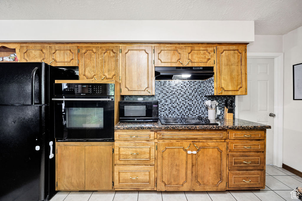 Kitchen with black appliances, under cabinet range hood, tasteful backsplash, brown cabinetry, and light tile patterned floors
