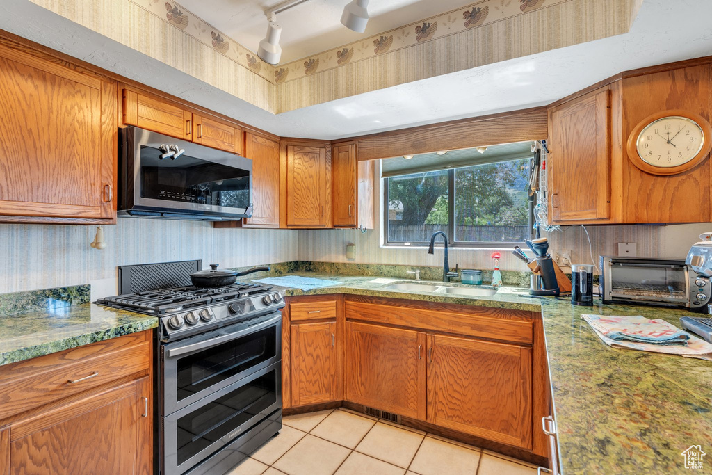 Kitchen featuring appliances with stainless steel finishes, brown cabinetry, light tile patterned floors, light stone countertops, and rail lighting