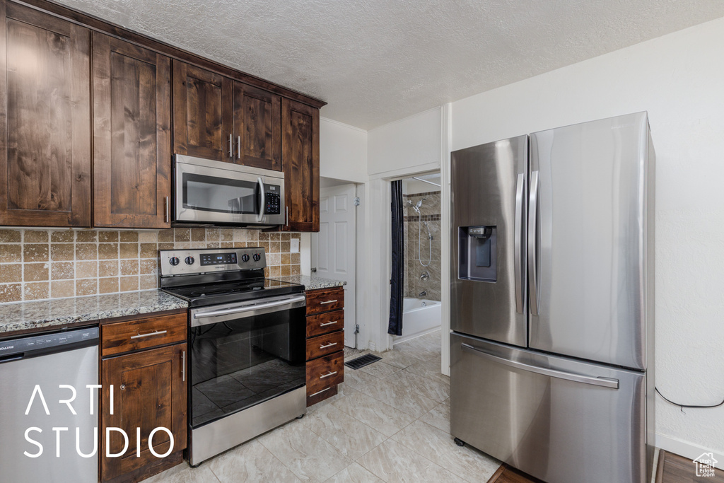 Kitchen with appliances with stainless steel finishes, decorative backsplash, dark brown cabinets, light stone counters, and a textured ceiling