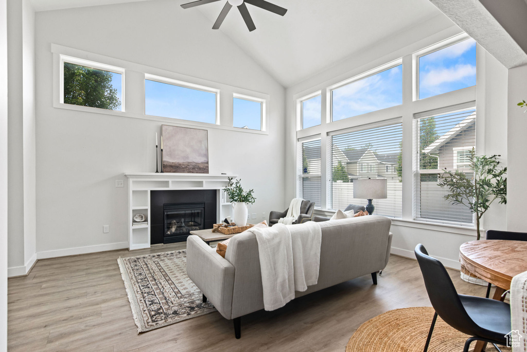 Living room with high vaulted ceiling, ceiling fan, healthy amount of natural light, a glass covered fireplace, and light wood-style flooring