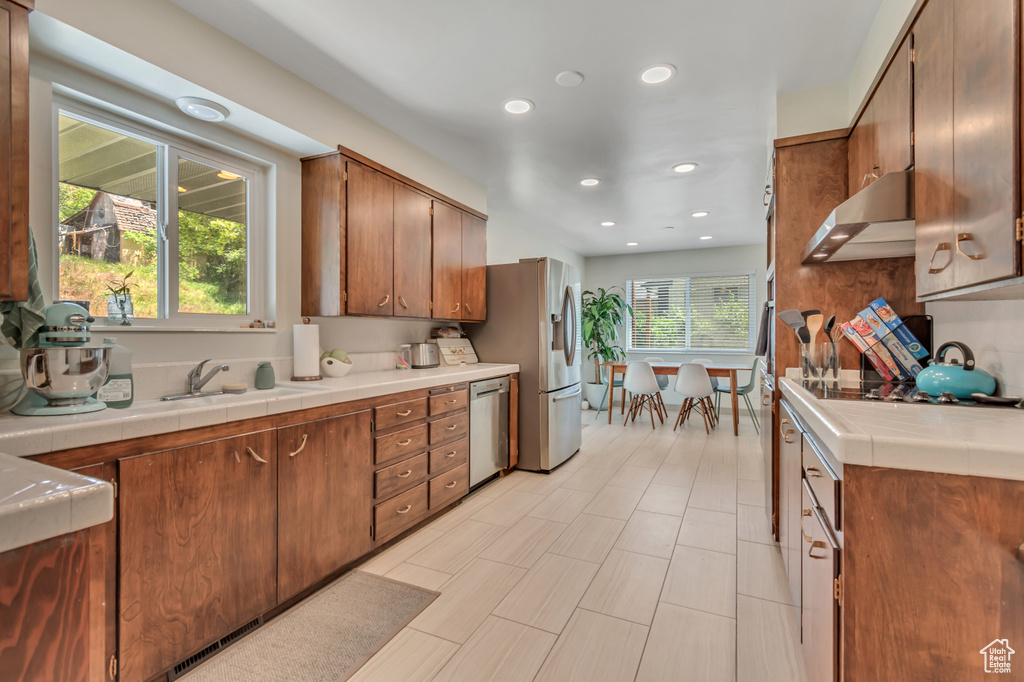 Kitchen featuring appliances with stainless steel finishes, under cabinet range hood, tile counters, recessed lighting, and brown cabinets