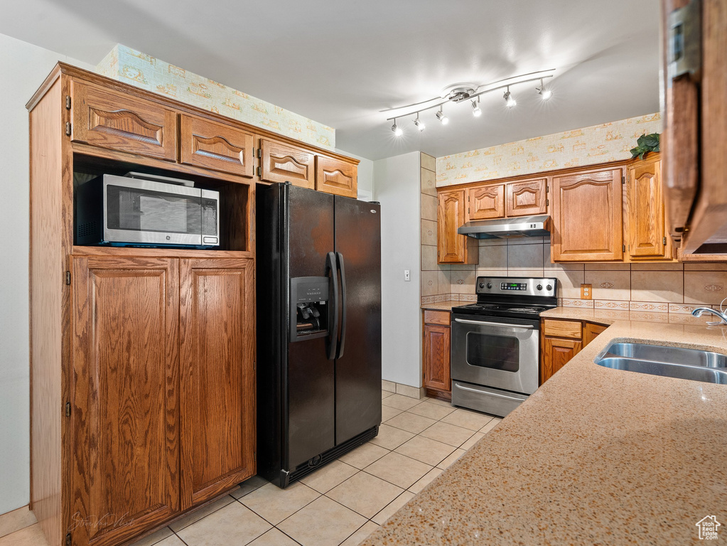 Kitchen featuring appliances with stainless steel finishes, under cabinet range hood, light tile patterned floors, light stone countertops, and brown cabinets