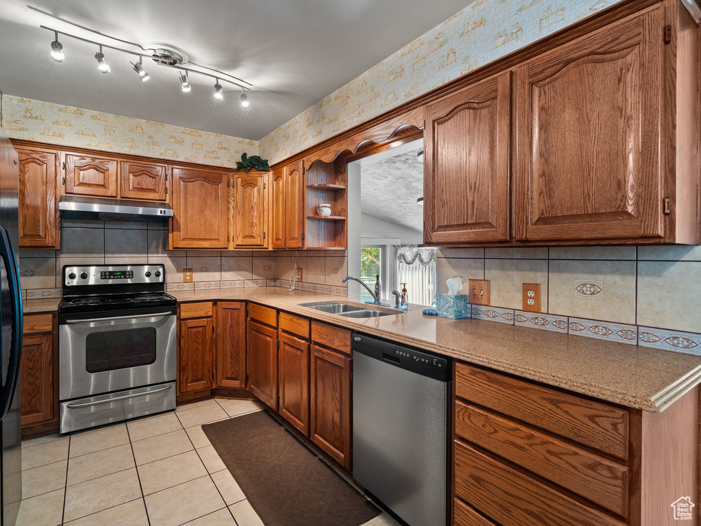 Kitchen with stainless steel appliances, wallpapered walls, under cabinet range hood, brown cabinetry, and light tile patterned floors