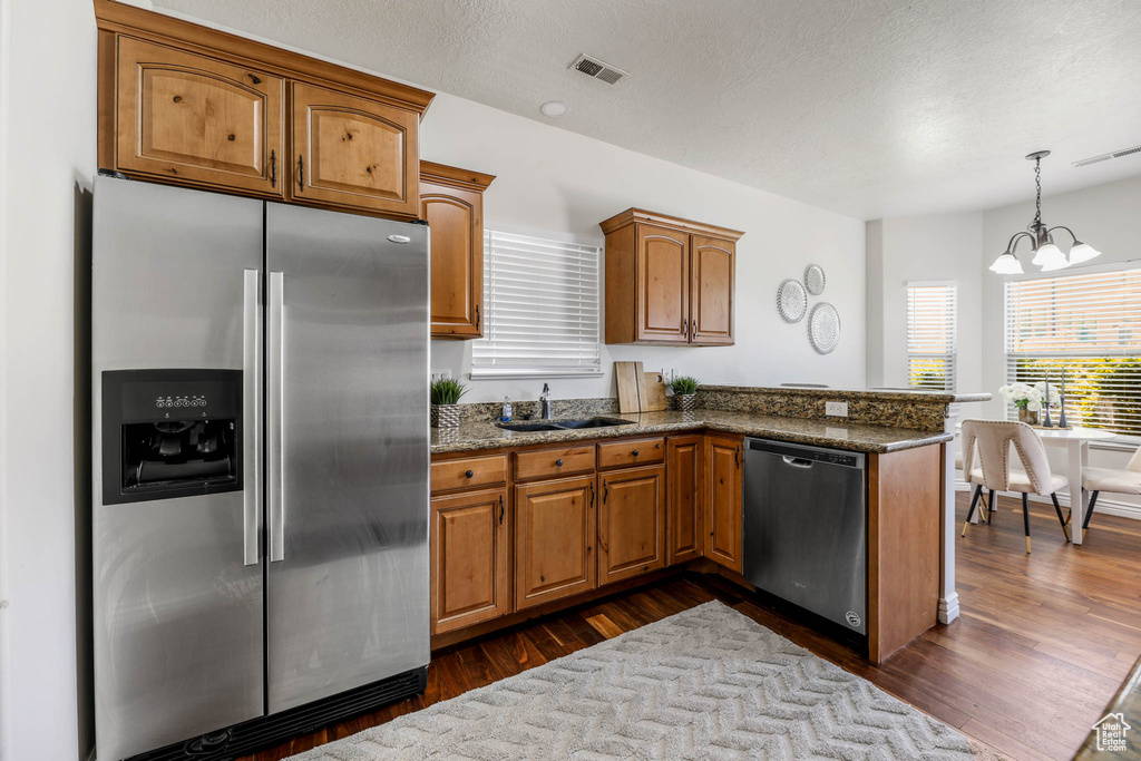 Kitchen with appliances with stainless steel finishes, a peninsula, a chandelier, brown cabinets, and dark stone countertops