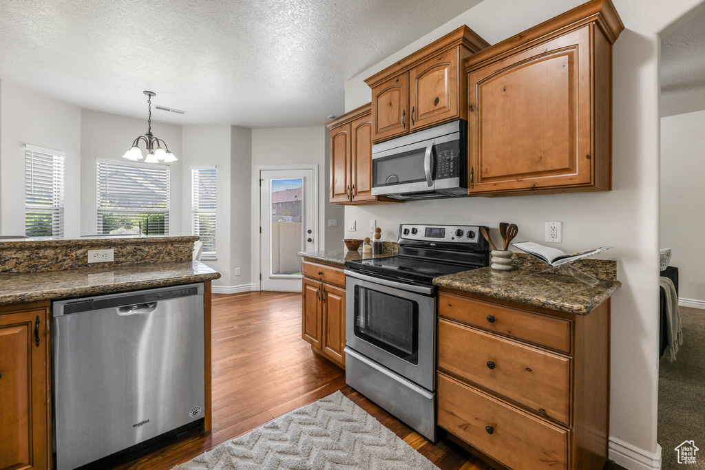 Kitchen with appliances with stainless steel finishes, dark countertops, brown cabinetry, dark wood-type flooring, and a textured ceiling