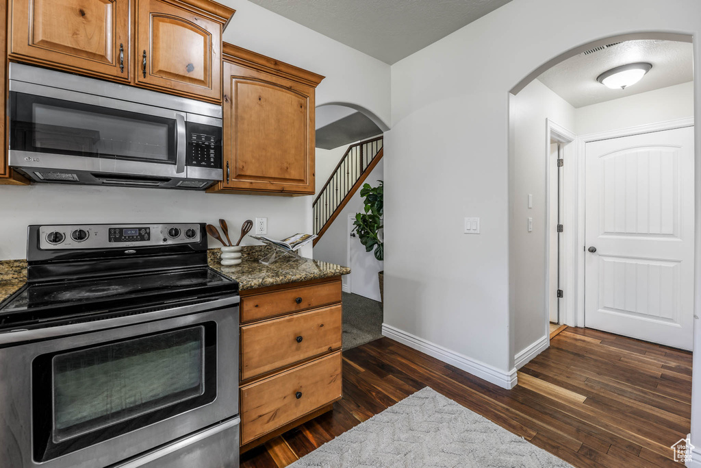 Kitchen featuring appliances with stainless steel finishes, arched walkways, dark wood-style floors, brown cabinetry, and dark stone countertops