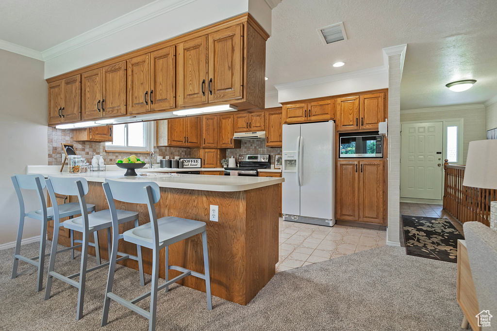Kitchen with white refrigerator with ice dispenser, stainless steel range with electric stovetop, a peninsula, brown cabinets, and crown molding