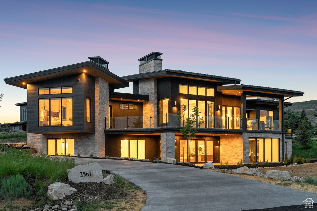 Back of property at dusk featuring a chimney, a balcony, stone siding, and driveway