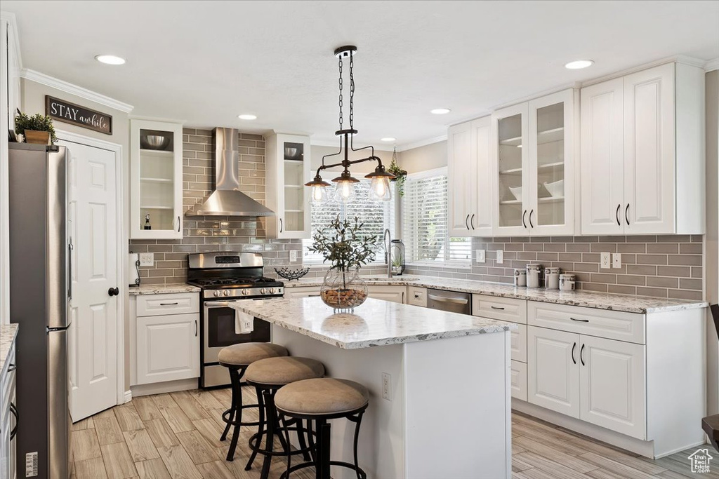 Kitchen with stainless steel appliances, wall chimney range hood, ornamental molding, white cabinets, and a center island