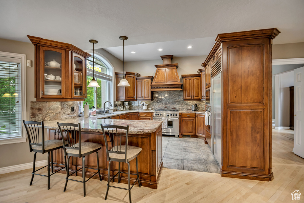 Kitchen with high end stove, a peninsula, brown cabinetry, custom range hood, and tasteful backsplash