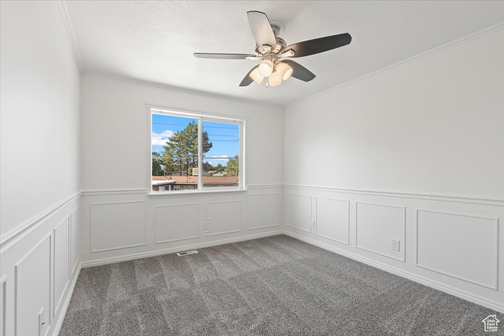 Carpeted empty room featuring crown molding, ceiling fan, a wainscoted wall, and a decorative wall