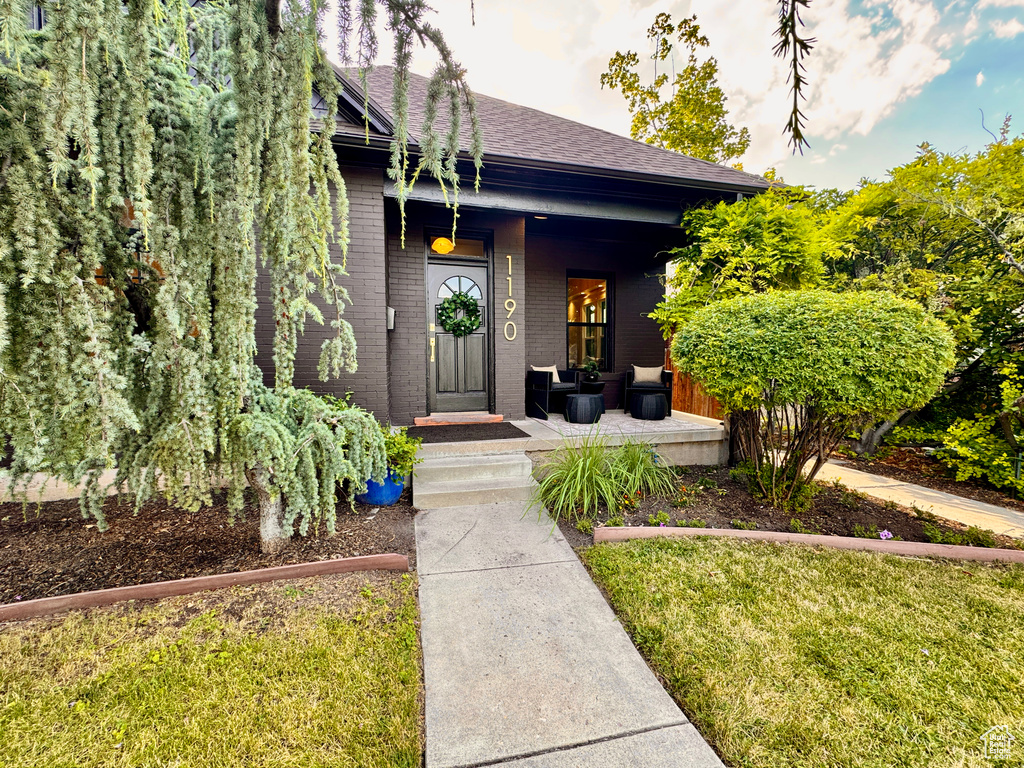 Doorway to property featuring brick siding, covered porch, a shingled roof, and a yard