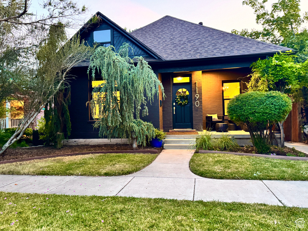 View of front of property featuring a shingled roof, a front yard, brick siding, and a porch