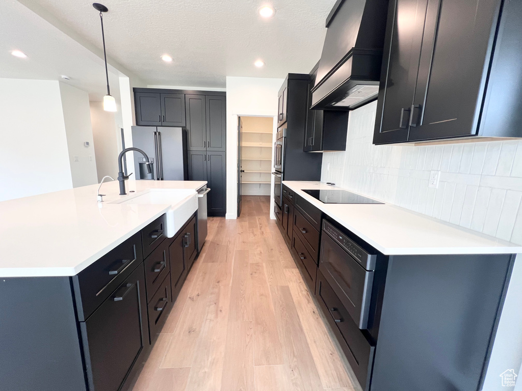 Kitchen with custom exhaust hood, appliances with stainless steel finishes, light wood-style flooring, tasteful backsplash, and light countertops