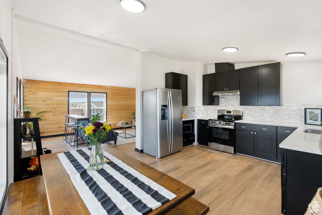 Kitchen featuring appliances with stainless steel finishes, under cabinet range hood, light countertops, dark cabinets, and wooden walls