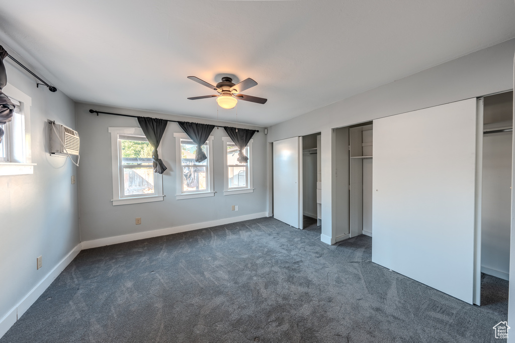 Unfurnished bedroom featuring a wall unit AC, dark carpet, multiple closets, and a ceiling fan