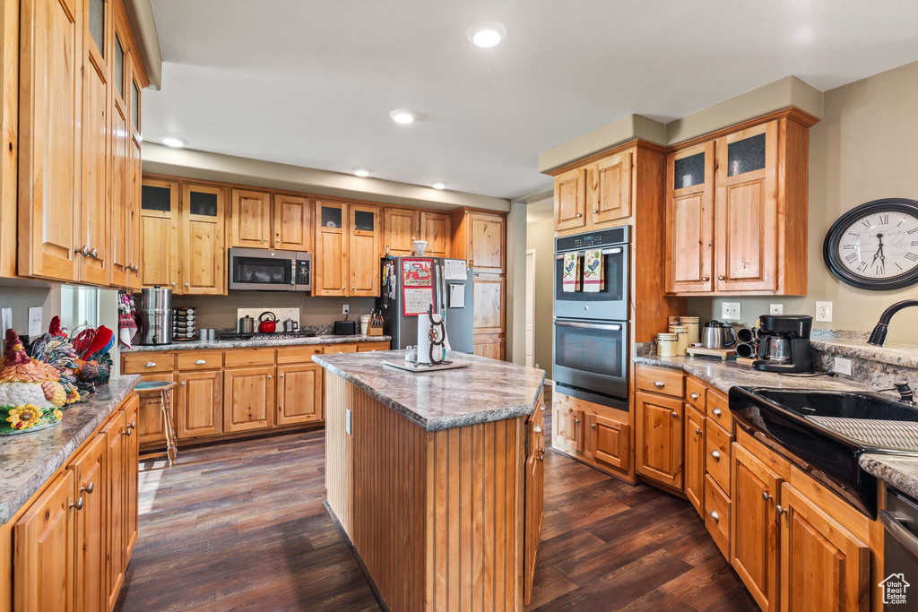 Kitchen with appliances with stainless steel finishes, dark wood finished floors, a center island, light stone countertops, and glass insert cabinets
