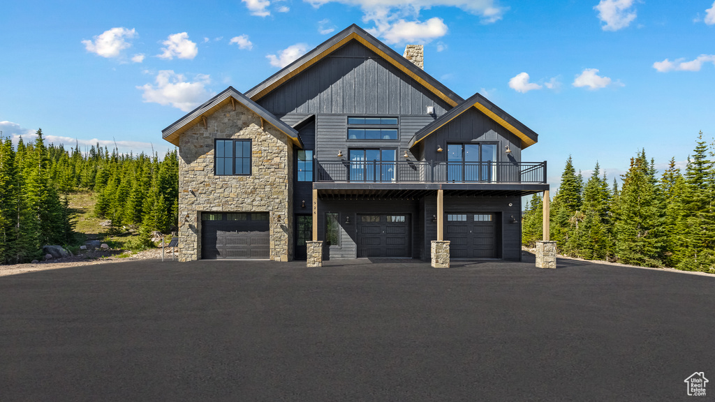 View of front of property featuring stone siding, a chimney, an attached garage, a balcony, and asphalt driveway