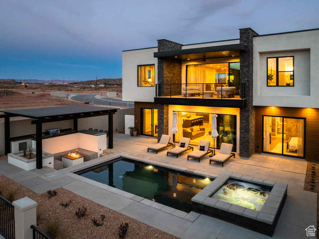 Back of house at dusk with a balcony, an in-ground hot tub, a fire pit, and stone siding